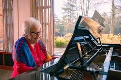 An elderly woman playing the piano by a sunlit window.