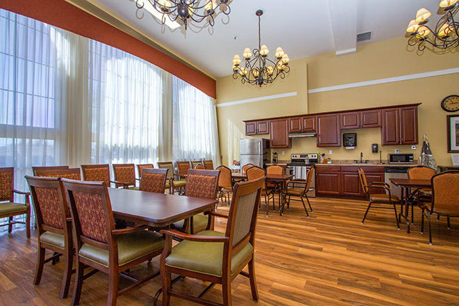 A spacious dining area with a kitchen, featuring a wooden floor, elegant chandeliers, and large windows with curtains.