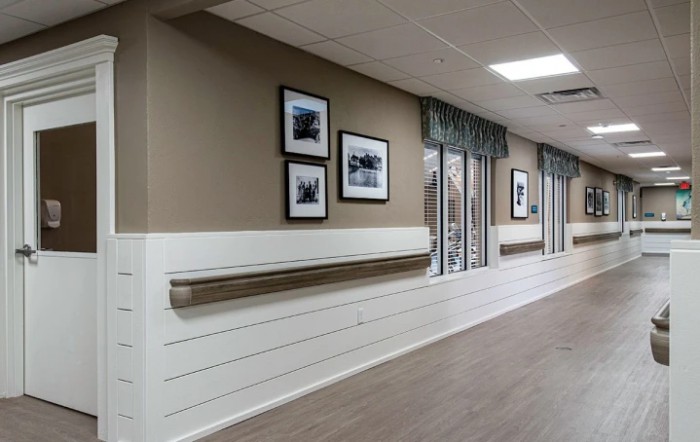 A well-lit hallway in a nursing home, featuring framed photographs and handrails along the walls.