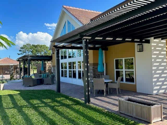 A house with a patio featuring a pergola and outdoor seating, surrounded by a green lawn under a clear blue sky.