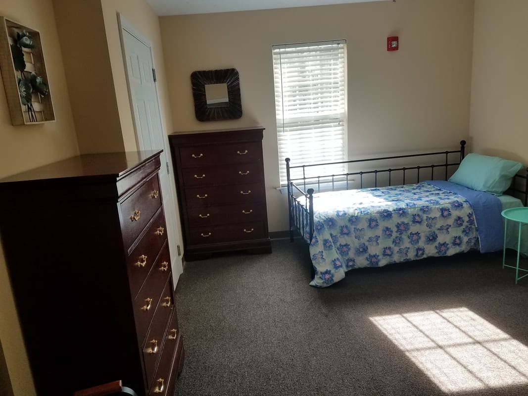 A bedroom with a single bed, two chests of drawers, and a window letting in sunlight.