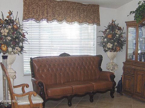 A traditional living room with a leather sofa, large window with blinds and curtains, and decorative floral arrangements.