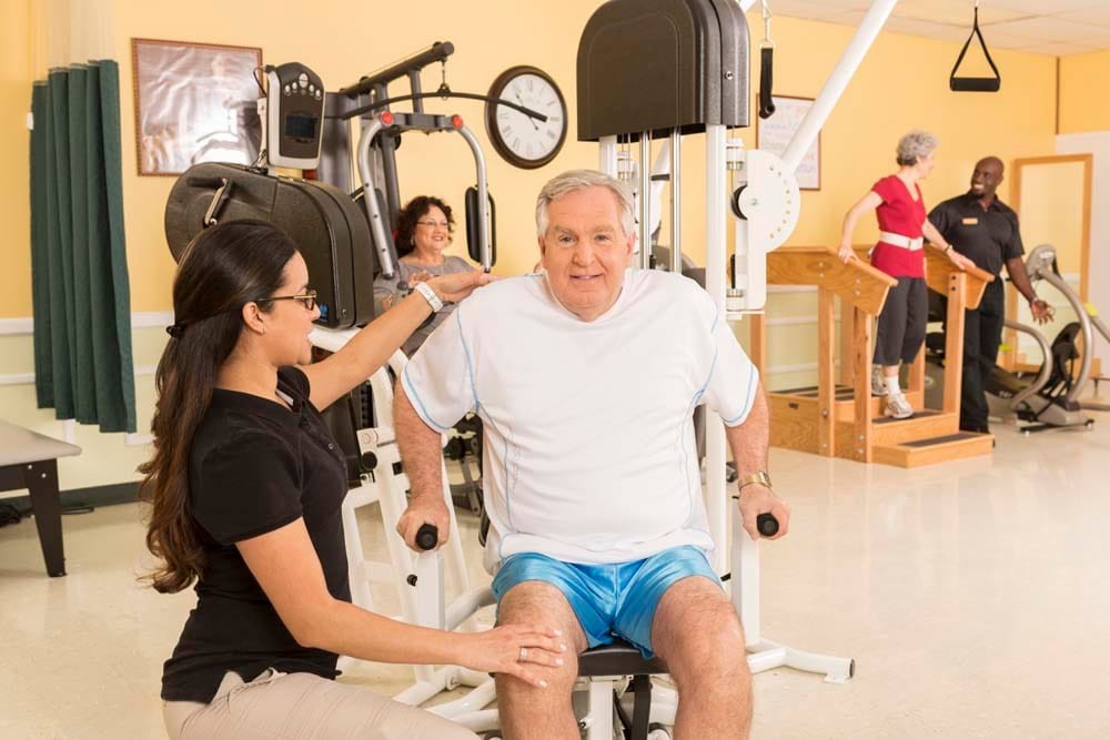 An elderly man receives guidance from a trainer while using exercise equipment in a gym.