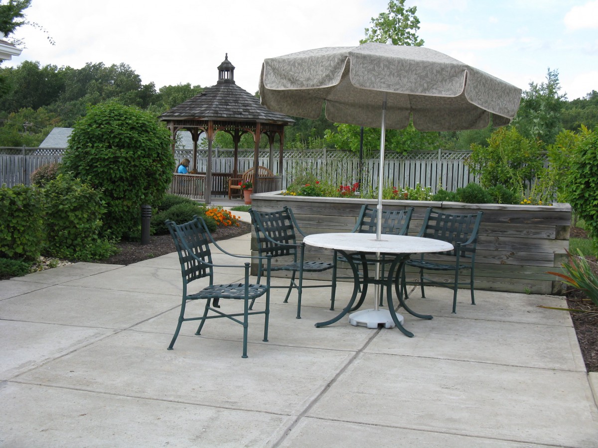 An outdoor patio with a table, chairs, and an umbrella set on a paved area, surrounded by greenery and a gazebo in the background.
