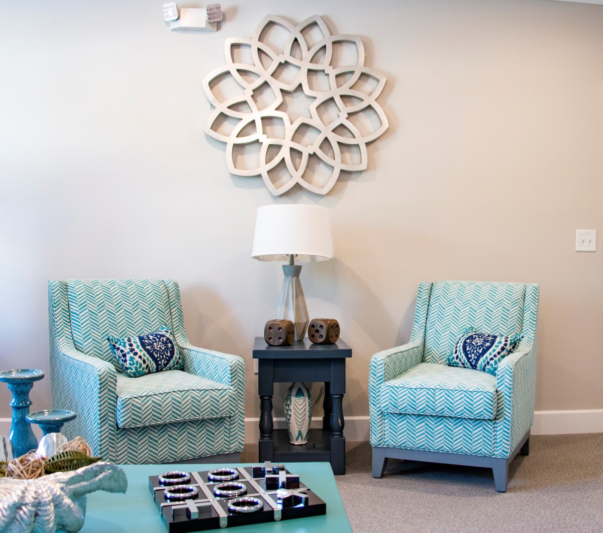 A cozy living room corner with teal armchairs, a side table holding a lamp, and decorative wall art.