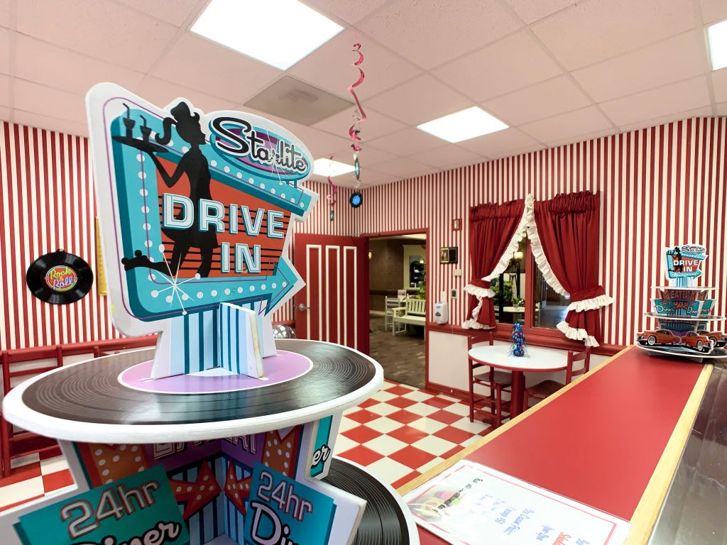 A retro-themed diner with red and white decor, featuring a 'Drive-In' sign and a classic checkerboard floor.