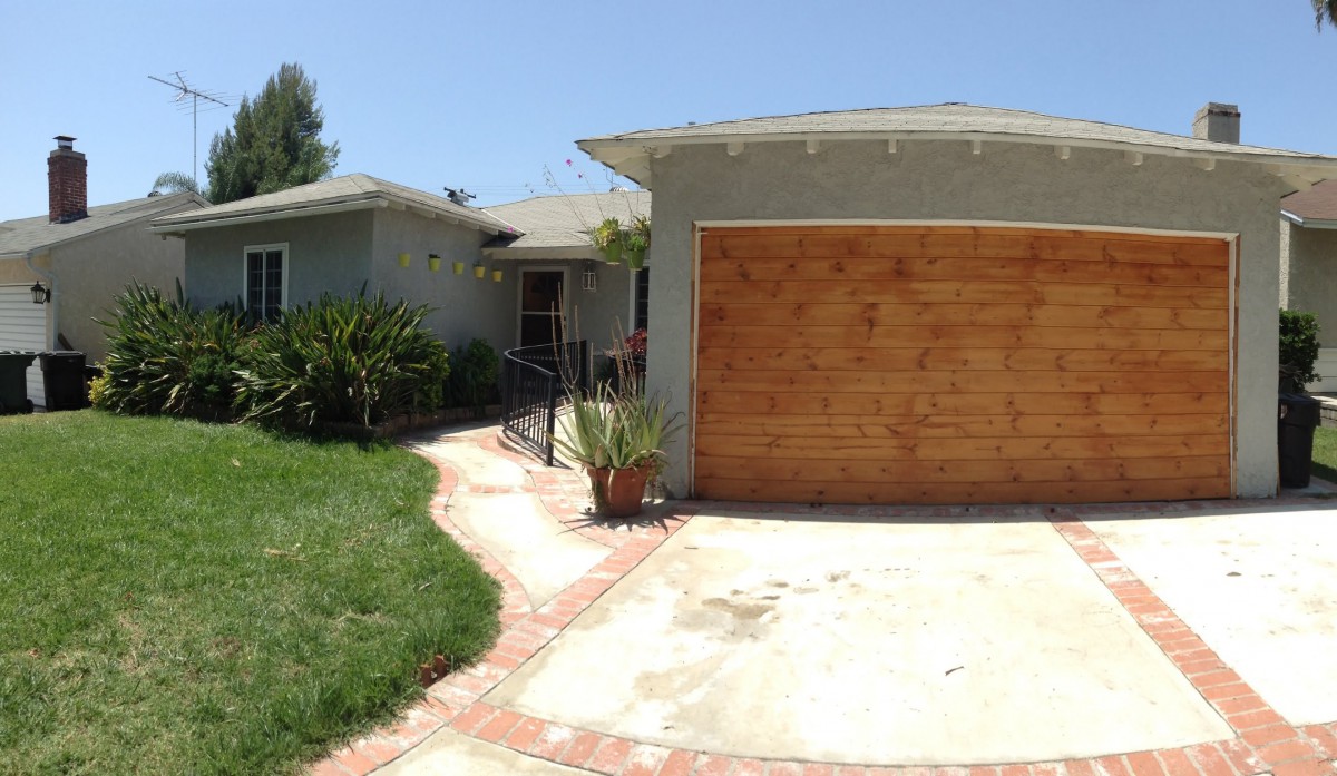 A suburban house with a wooden garage door and a driveway, surrounded by greenery under a clear blue sky.