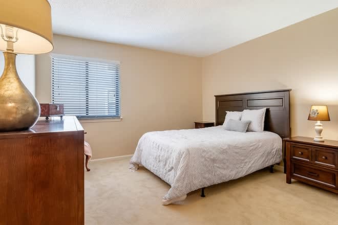 A minimalist bedroom with a neatly made bed, wooden furniture, and a window with blinds.