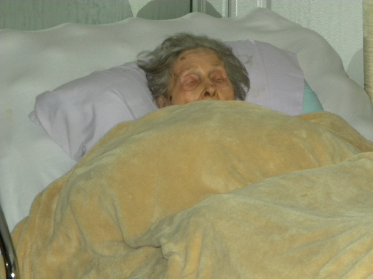 An elderly person is resting in bed, covered with a blanket.