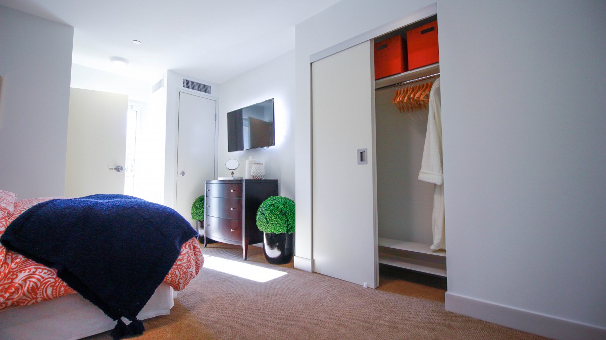 A bright and modern bedroom with a television, dresser, and an open closet displaying robes. The room is decorated with potted plants.