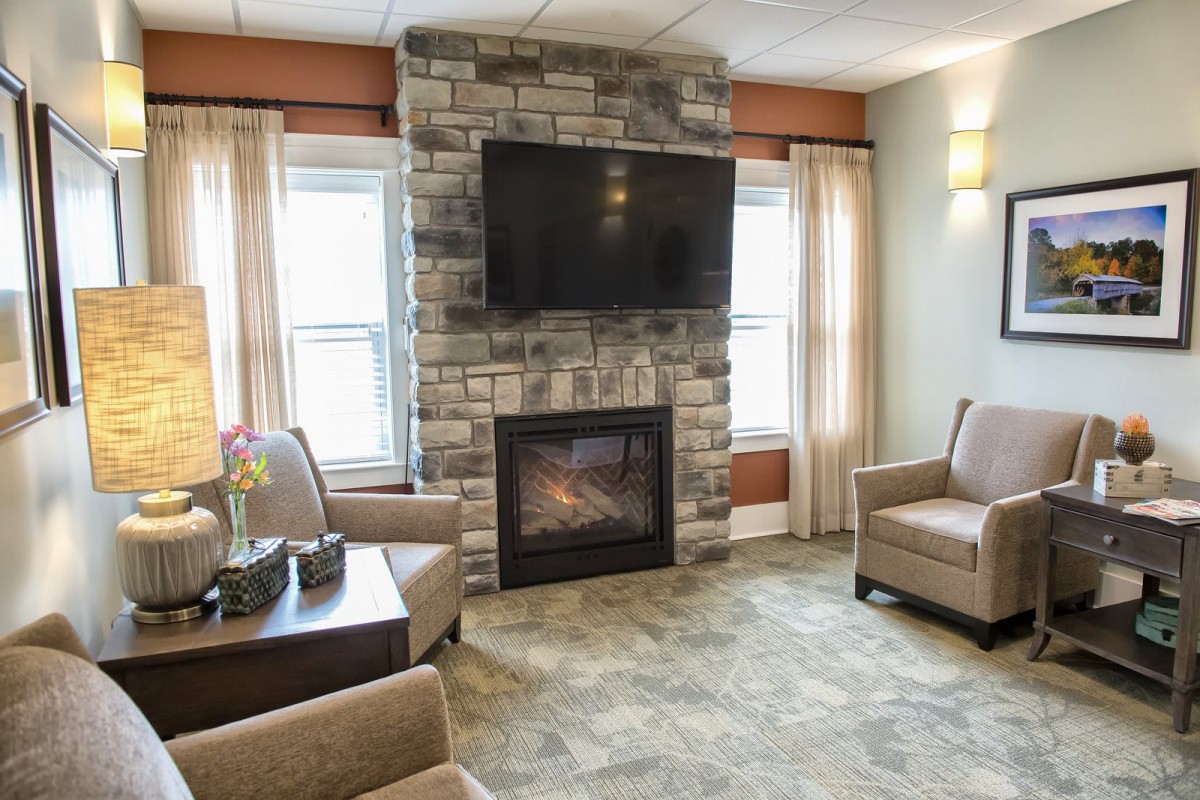 A cozy living room featuring a stone fireplace with a television mounted above, surrounded by comfortable armchairs and large windows.