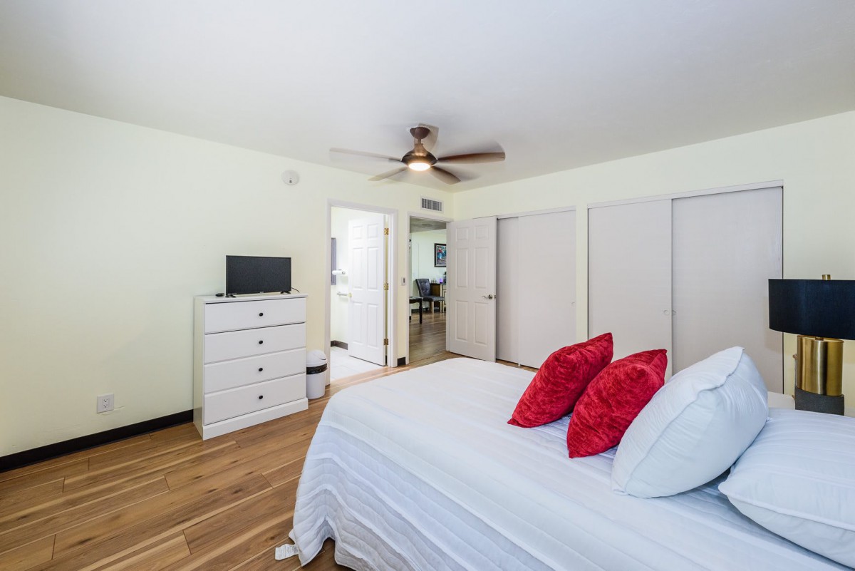 A cozy bedroom featuring a bed with red pillows, white linens, a white dresser, and a ceiling fan. The room is decorated with hardwood floors and neutral walls.