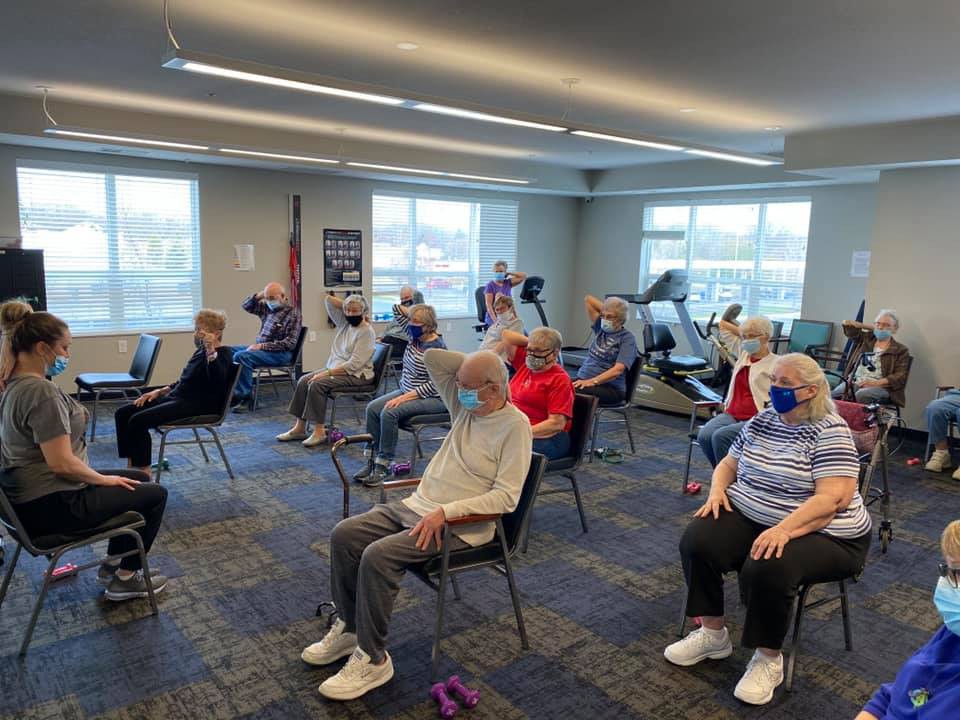 A group of seniors participating in an exercise class while seated in chairs, all wearing masks inside a community center.