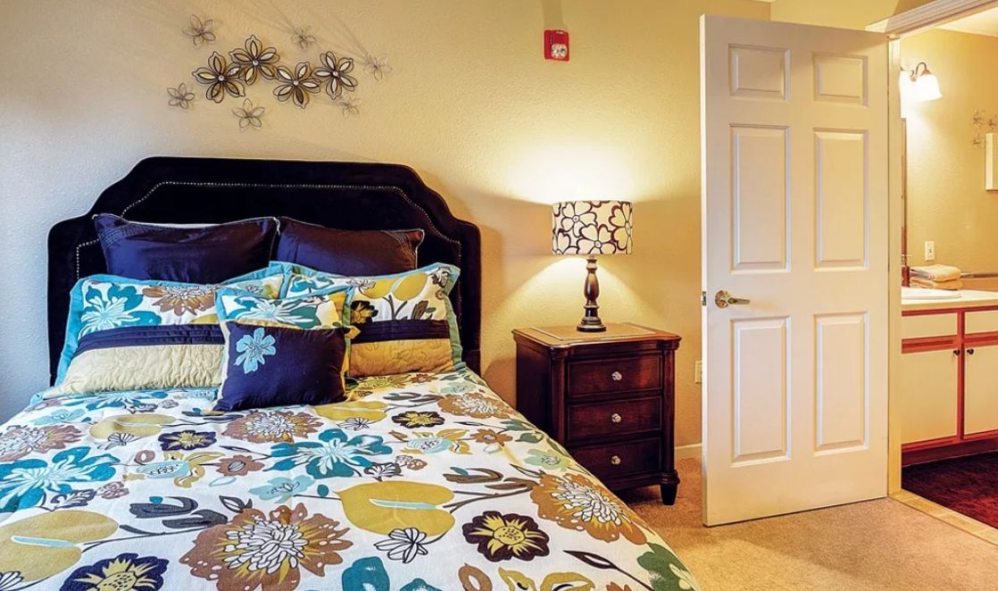 A cozy bedroom featuring a bed with floral-patterned bedding and decorative pillows, next to a nightstand with a lamp.