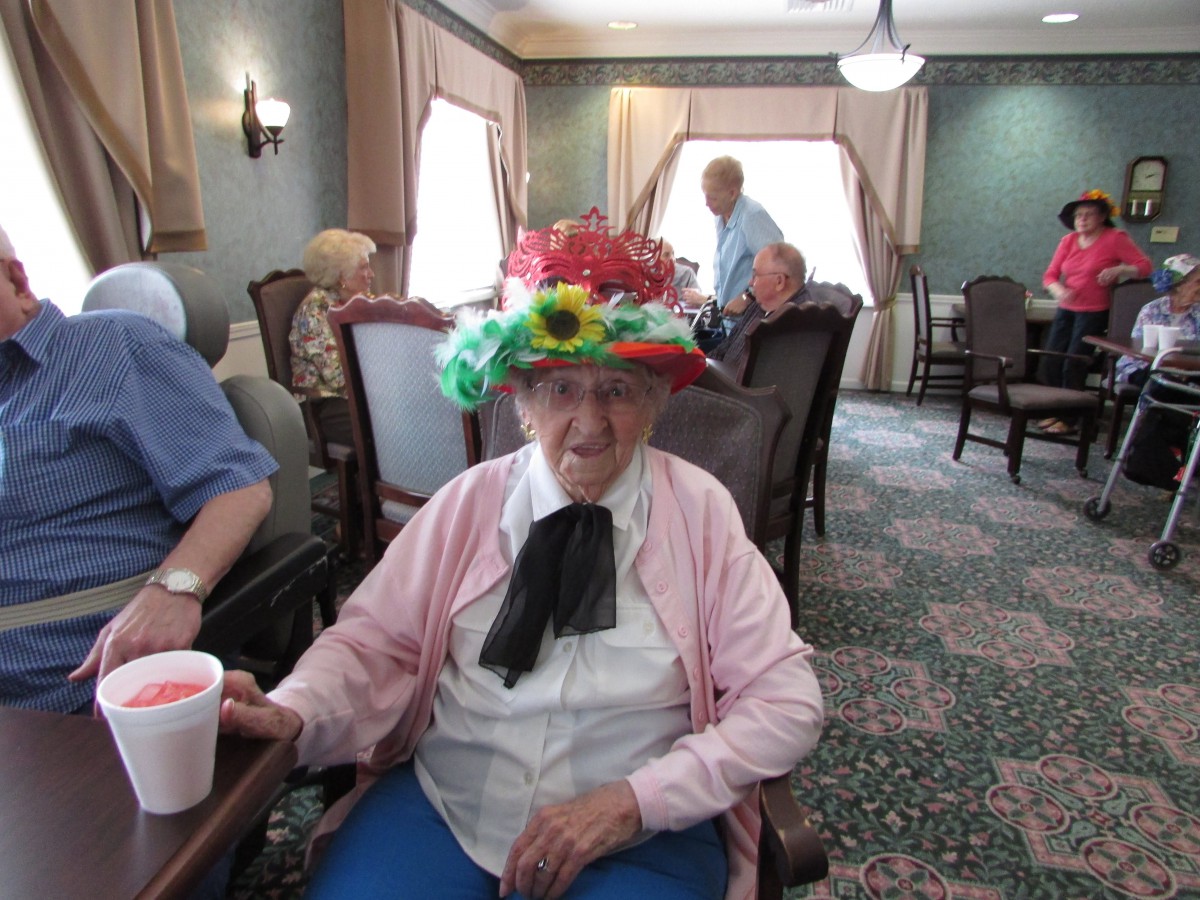 An elderly person wearing a colorful hat sits in a room during a social gathering.