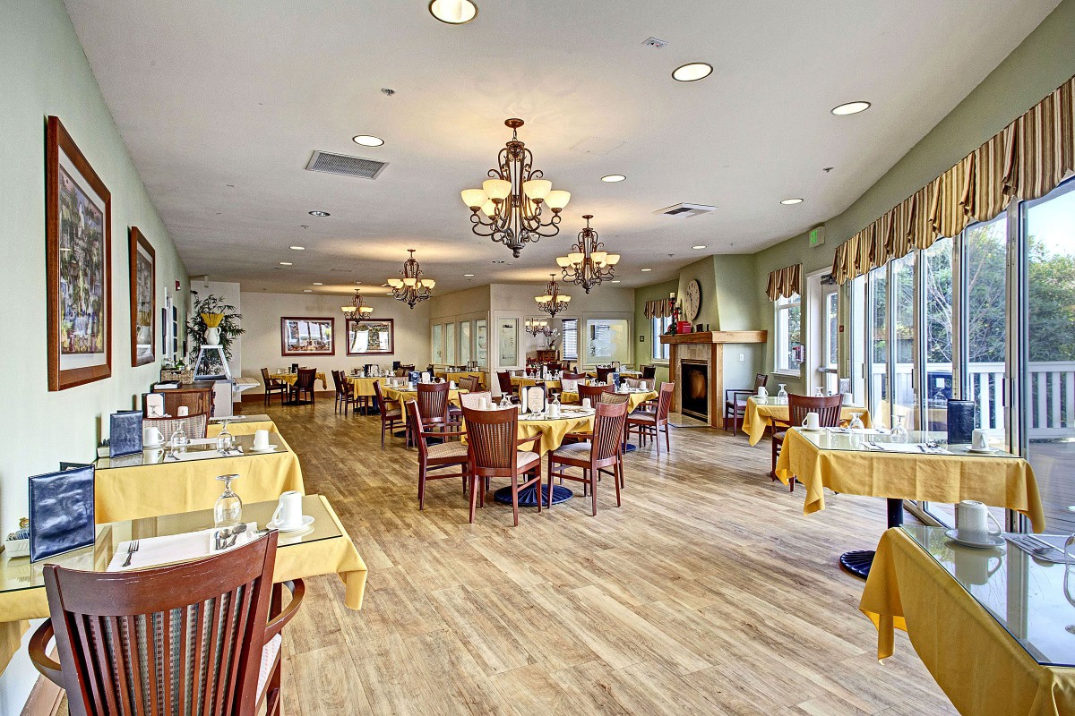 A spacious, elegantly decorated dining room featuring wooden floors, chandeliers, and neatly arranged tables with yellow tablecloths.
