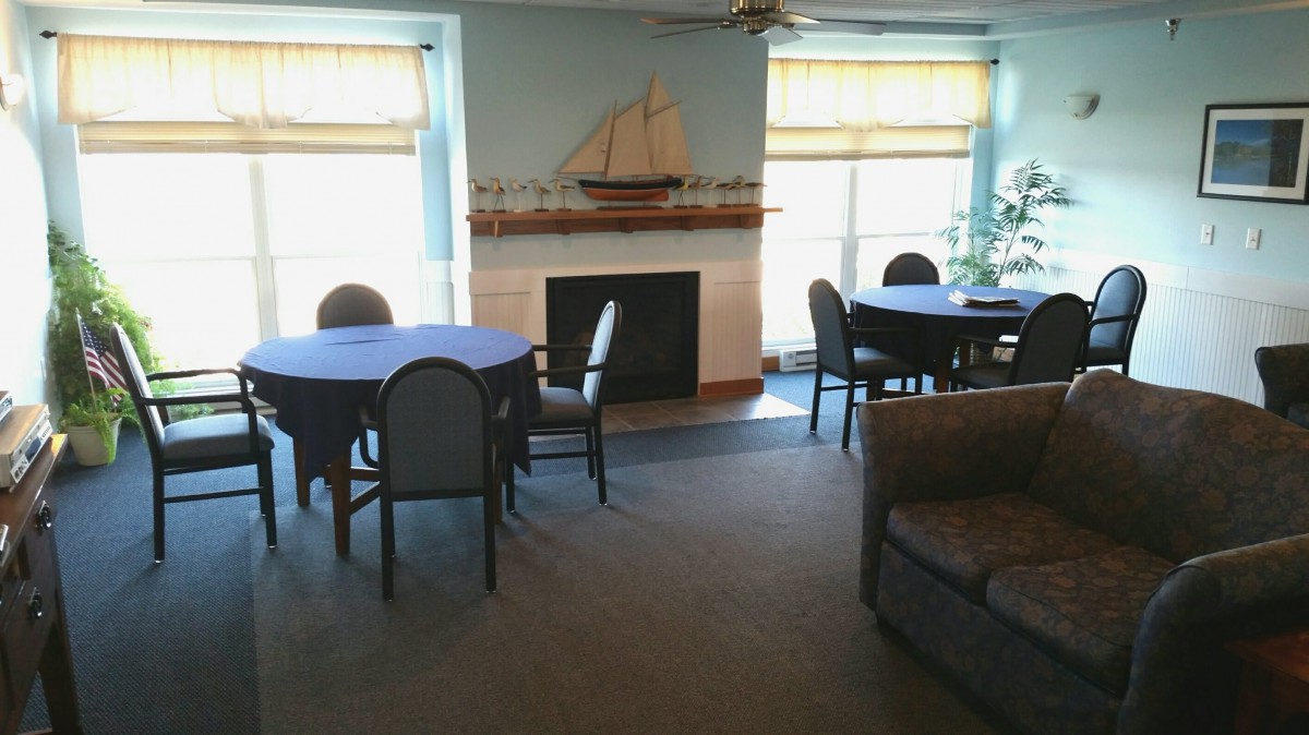 A cozy living room with tables and chairs arranged around a fireplace, decorated with a model ship on the mantel.