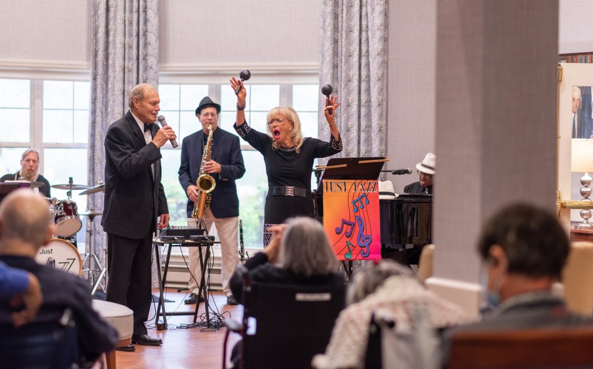 A band performs indoors with a singer, saxophonist, and pianist entertaining an audience.