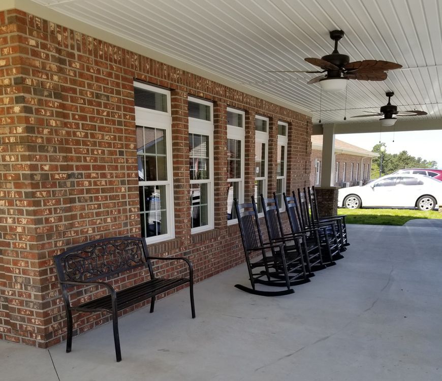 A porch with multiple rocking chairs, a bench, and a ceiling fan along a brick wall.