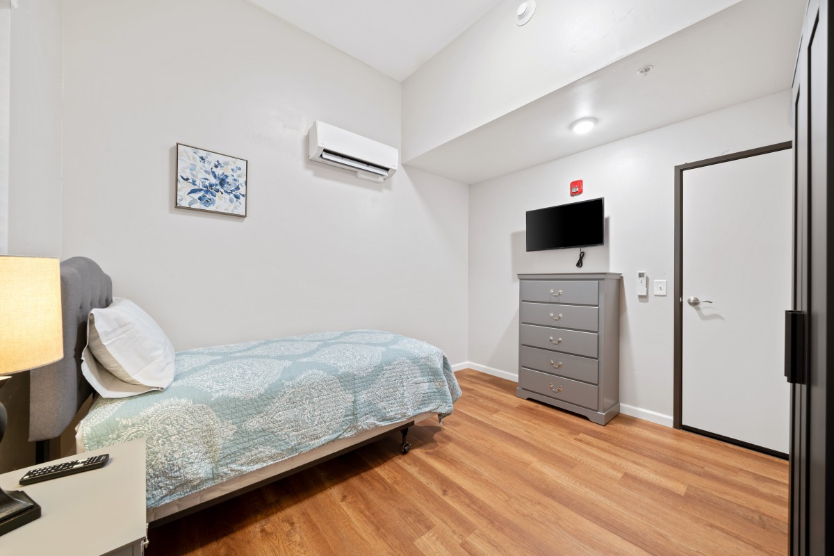 A minimalist bedroom with wooden flooring, featuring a single bed, dresser, mounted television, and air conditioner.