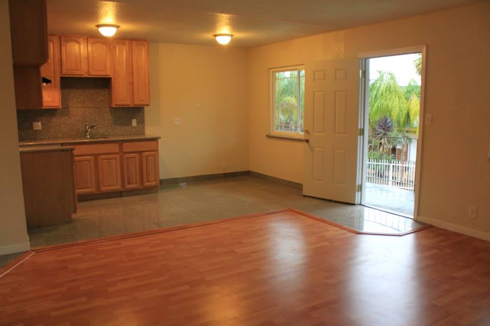 An empty room featuring a small kitchen area with wooden cabinets, an open door leading outside, and a window allowing natural light.