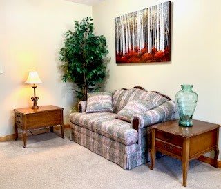 A cozy living room corner with a patterned sofa, surrounded by side tables, a decorative lamp, and an indoor plant, featuring a colorful forest painting.
