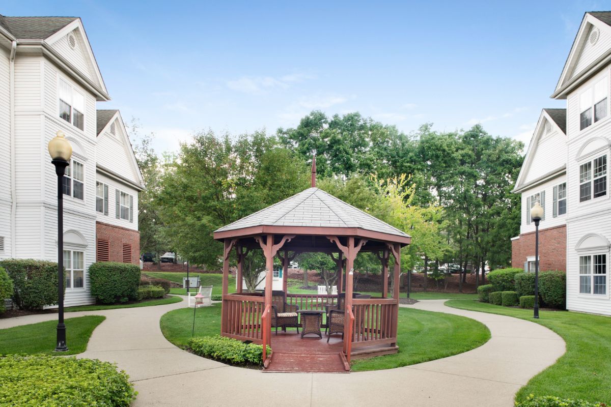 A charming wooden gazebo surrounded by a lush green lawn and pathway, set against a backdrop of trees and a residential building.