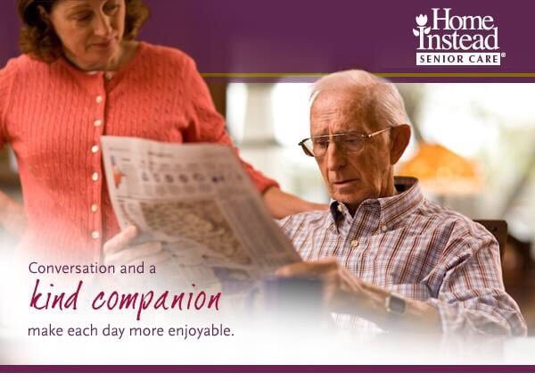 An elderly man reading a newspaper with a caregiver providing companionship at home.