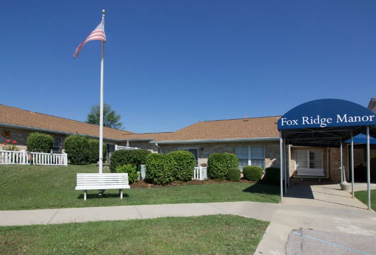 A building with an American flag, surrounded by grass and bushes, featuring a bench and an awning at the entrance.