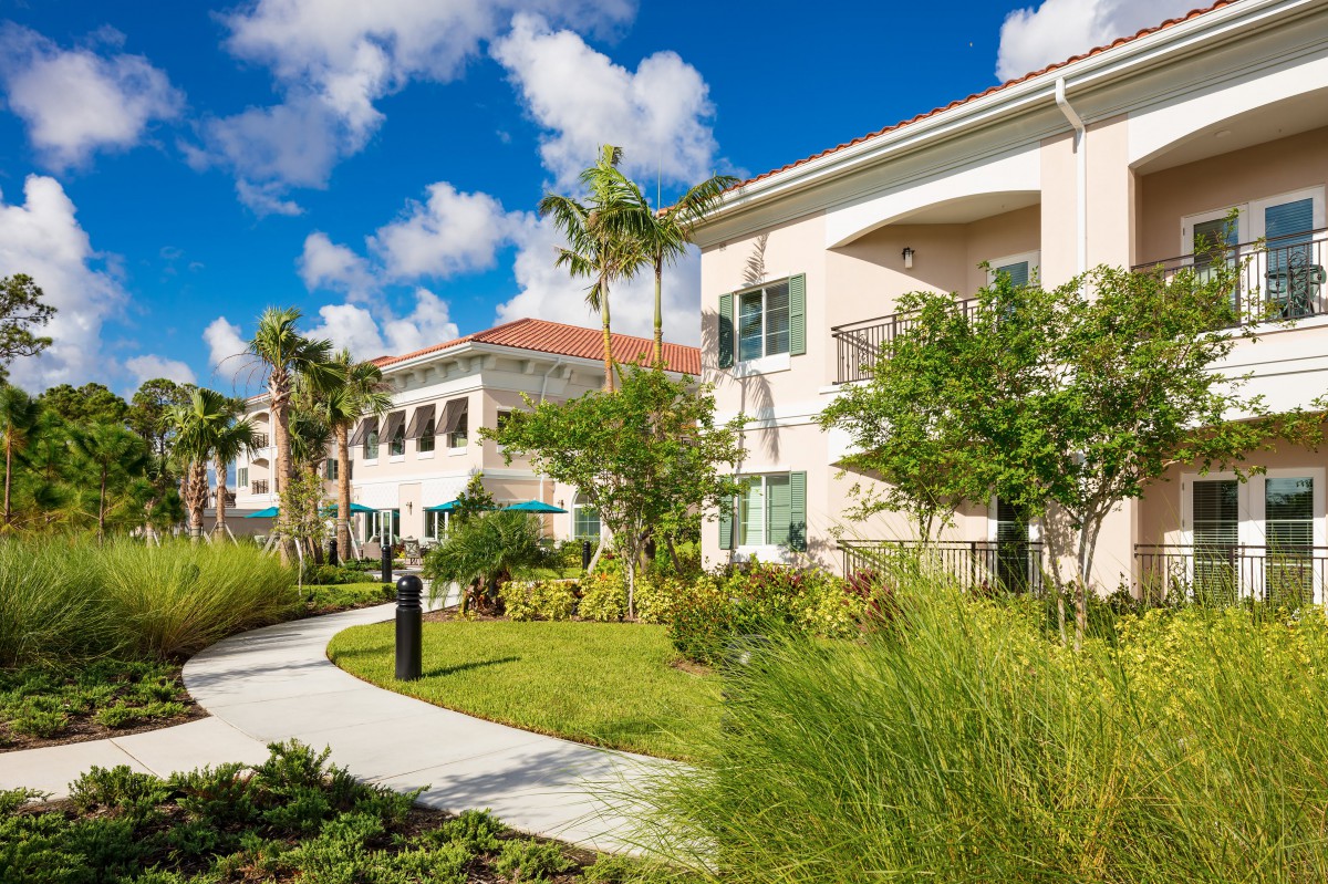 A tropical residential building surrounded by lush landscaping and palm trees under a bright blue sky.