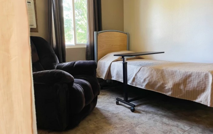 A cozy bedroom with a single bed, recliner, and adjustable side table beside a window.