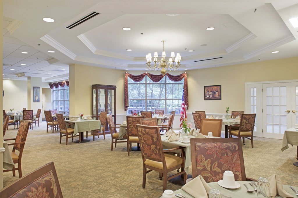 A well-lit dining room featuring elegantly set tables and chairs with a chandelier and large windows adorned with curtains.