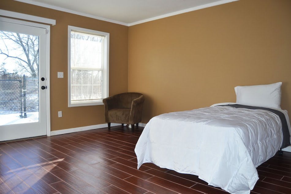 A spacious bedroom with hardwood flooring, featuring a bed, chair, and large window.