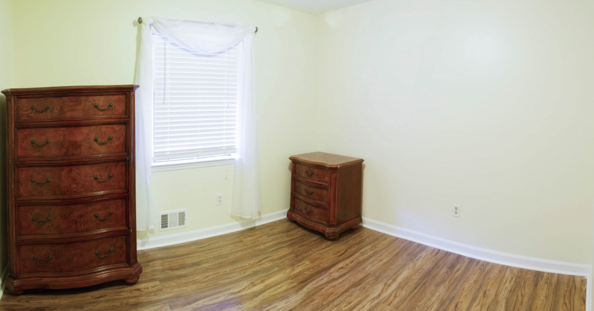 An empty room with wooden flooring, a small dresser, and a window with blinds.