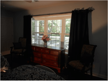 A cozy bedroom featuring a dresser with flowers, flanked by chairs and framed by dark curtains and a window.