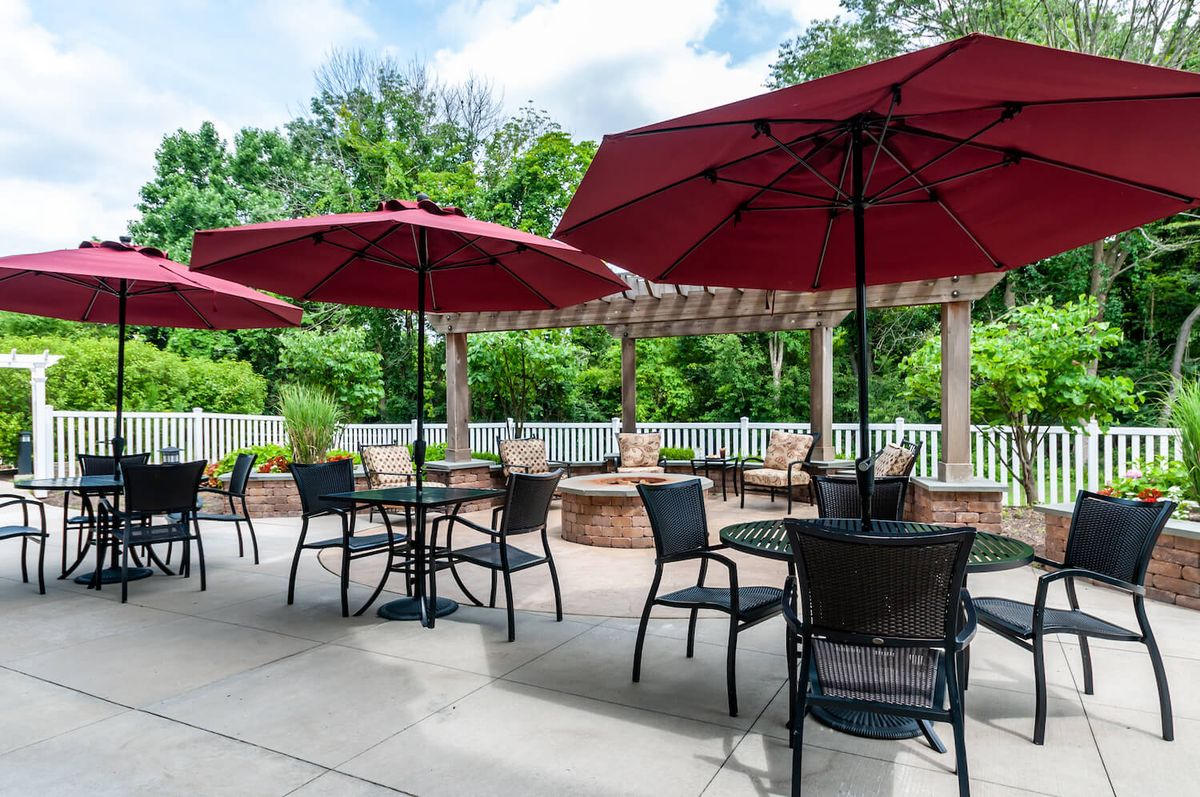An inviting outdoor patio area with tables, chairs, and red umbrellas set against a lush green background.