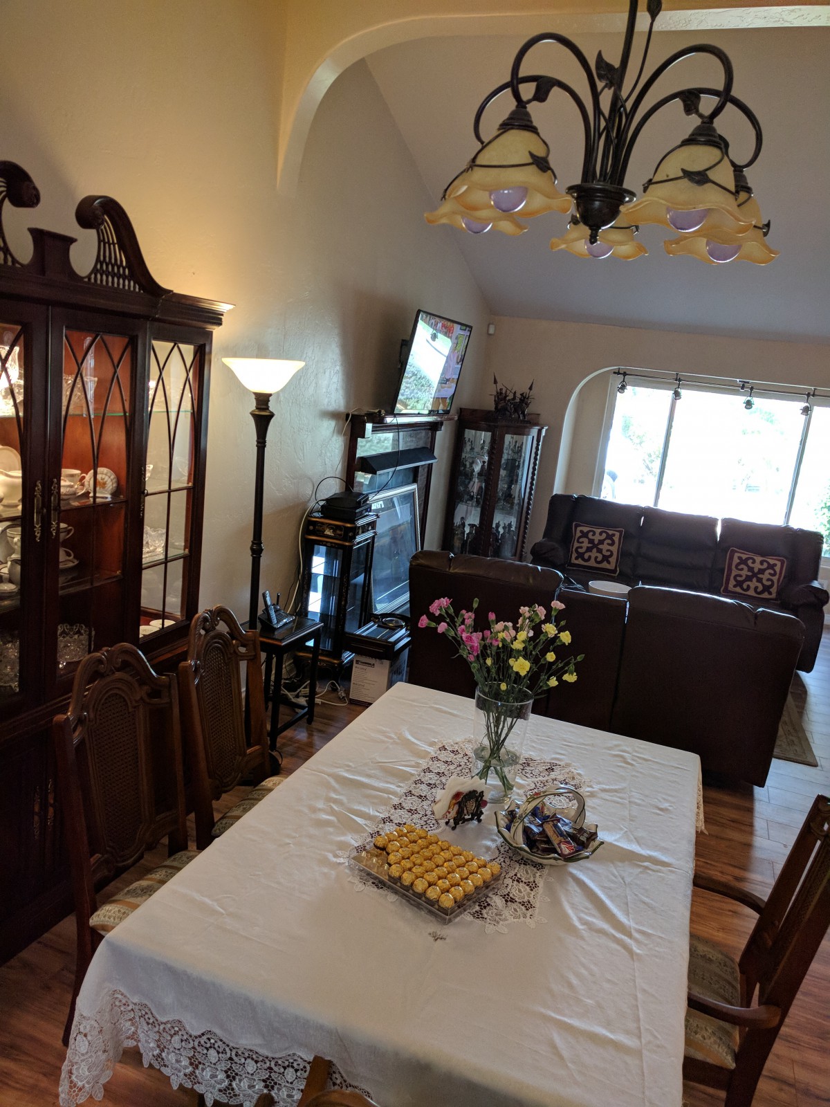 A cozy dining and living area with a table set for entertaining, featuring flowers, comfortable seating, and a television.