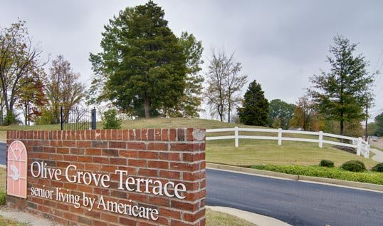 A brick signpost in front of a landscaped area with trees and a white fence.