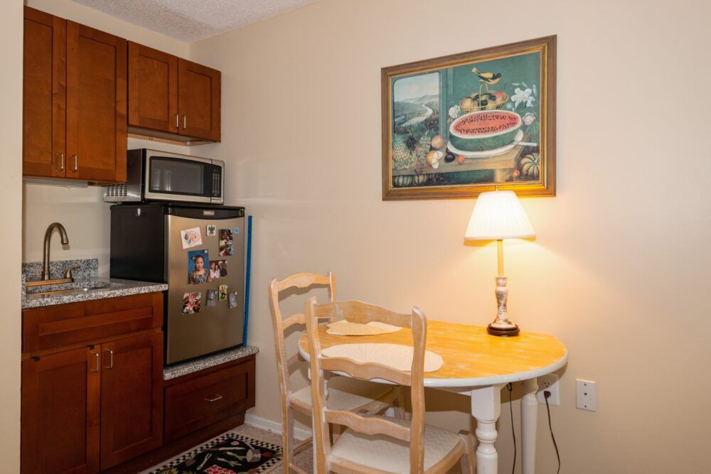 A cozy kitchen corner with a small dining table and chairs, illuminated by a table lamp. A painting hangs on the wall above the warm wooden cabinetry.