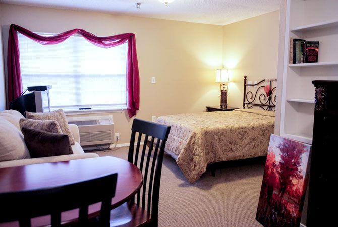 A cozy bedroom with a bed, table, chairs, and a lamp near a window with red curtains.