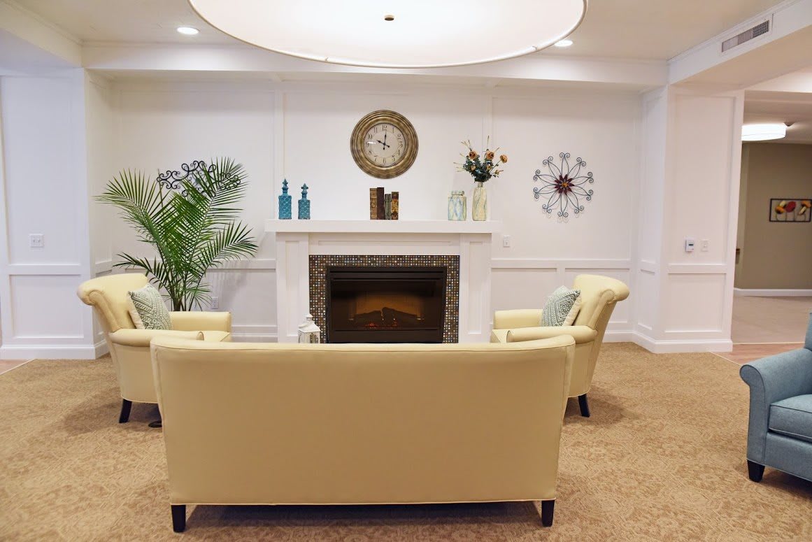 A cozy living room with plush beige sofas facing a white fireplace, adorned with decorative items and a wall clock.