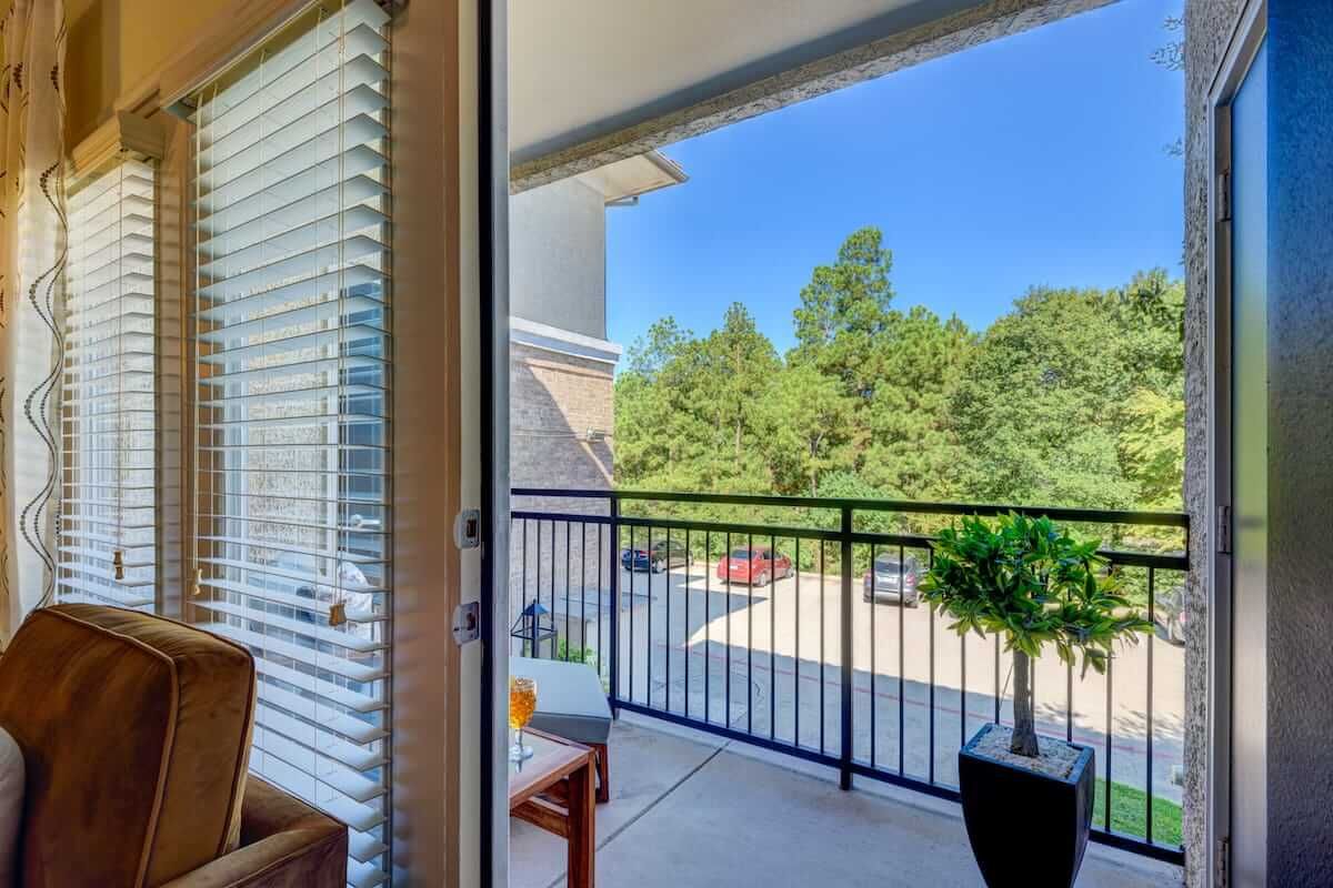A cozy balcony with outdoor seating overlooking a view of trees under a clear, sunny sky.