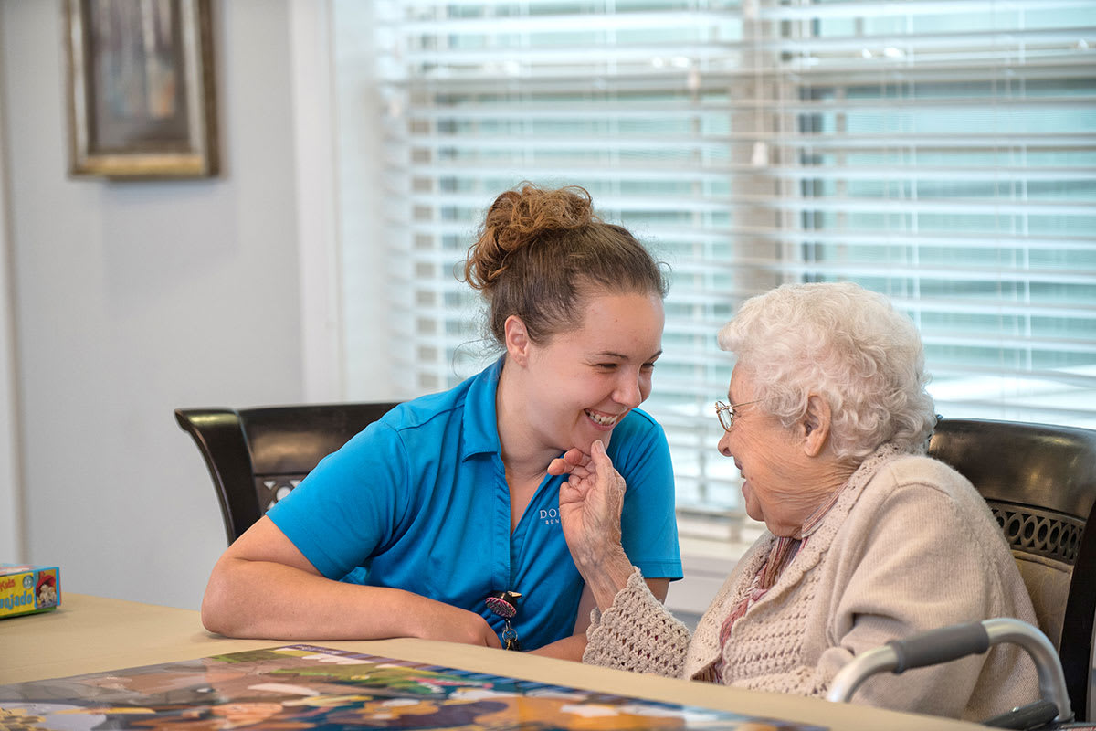 A caregiver and a senior woman share a warm moment of conversation indoors.