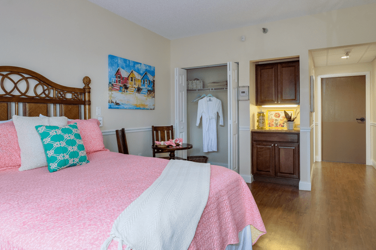 A cozy bedroom with a pink bedspread, artwork on the wall, and a small kitchenette.