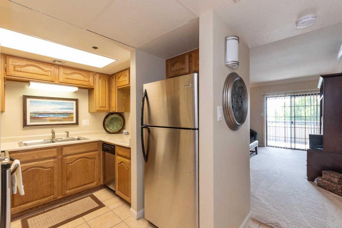 A modern apartment kitchen with wooden cabinets and a stainless steel fridge adjacent to an open living area.