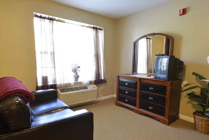 A cozy bedroom featuring a wooden dresser with an attached mirror, a TV on top, and natural light filtering through the window.