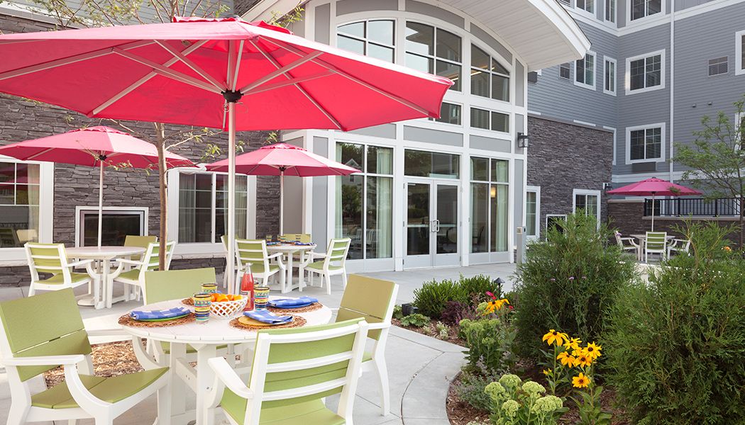 A cozy outdoor patio area with tables, chairs, and red umbrellas surrounded by a garden.