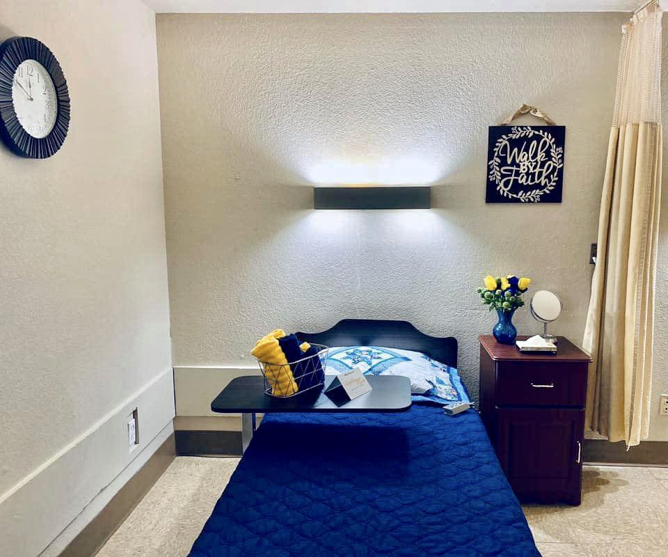 A neatly arranged hospital room with a single bed, overbed table, and nightstand adorned with flowers and wall decor.