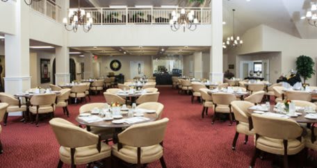 A spacious dining area with round tables and cushioned chairs, featuring elegant chandeliers and a red carpeted floor.