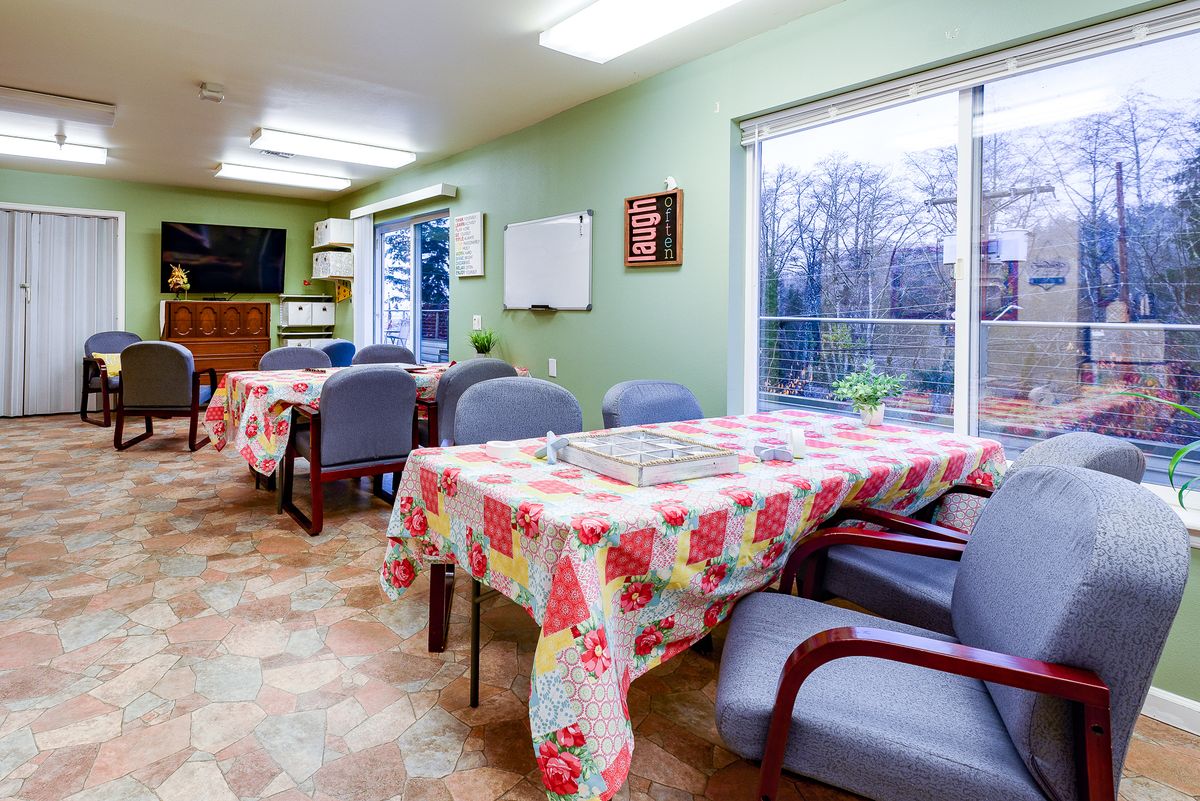 A cozy dining area with tables covered in floral tablecloths, surrounded by chairs and illuminated by bright ceiling lights.